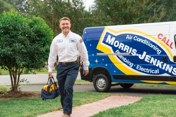 A technician in uniform carries a tool bag while walking toward a house, with a Morris-Jenkins service van parked in the background, ready to provide expert indoor air quality services.