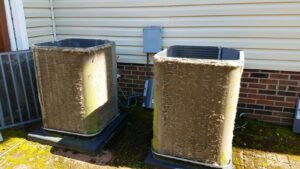 Two outdoor HVAC units with heavily dirt-clogged coils stand on pads next to a house with beige siding and brick trim, highlighting the need for maintenance and professional indoor air quality services.