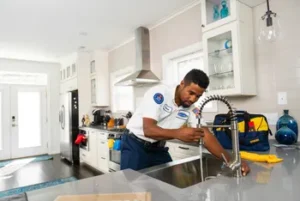 A plumber in uniform repairs a kitchen faucet at a modern home. Tools and a tool bag are on the counter next to him.