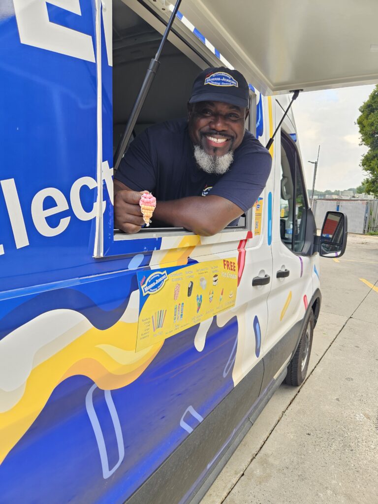 A man in a uniform leans out of an ice cream truck window, smiling and holding a soft serve cone.