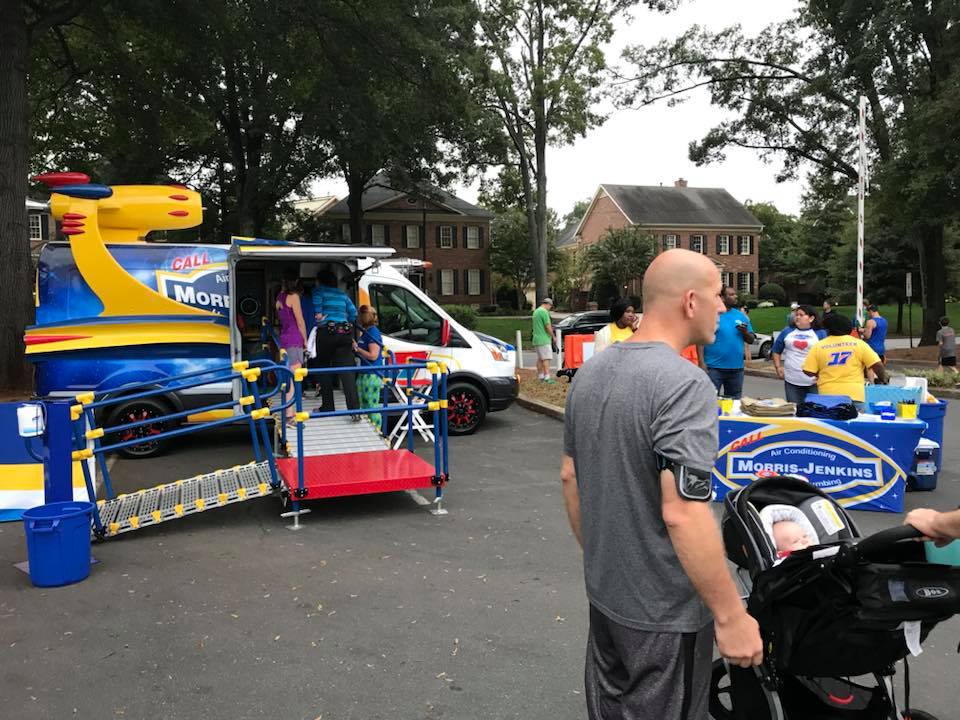 People gather near a Morris-Jenkins van and promotional booth in a suburban neighborhood, with children and adults participating in outdoor activities.
