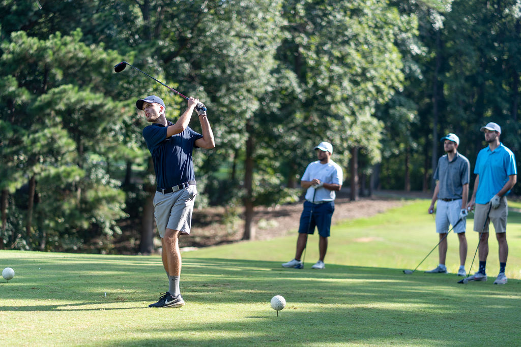 A man swings a golf club on a green golf course while three other men stand in the background watching.