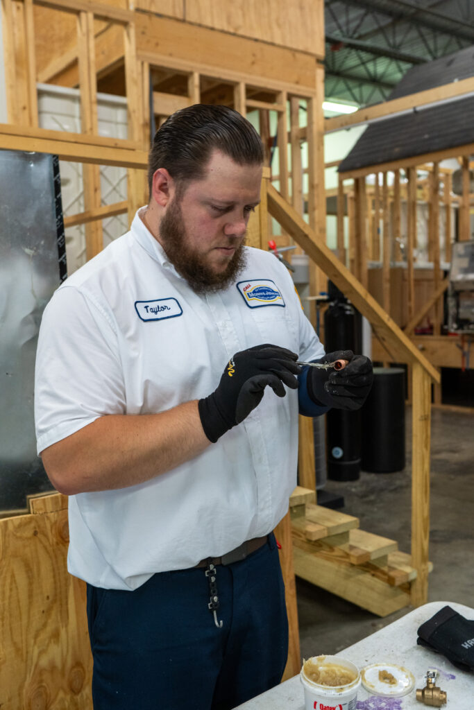 A man in a work uniform and gloves applies material to a pipe fitting in a workshop with wooden structures in the background.