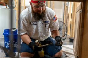 A technician wearing gloves and safety gear uses a handheld torch while kneeling on the floor in a workshop with exposed wooden framing.