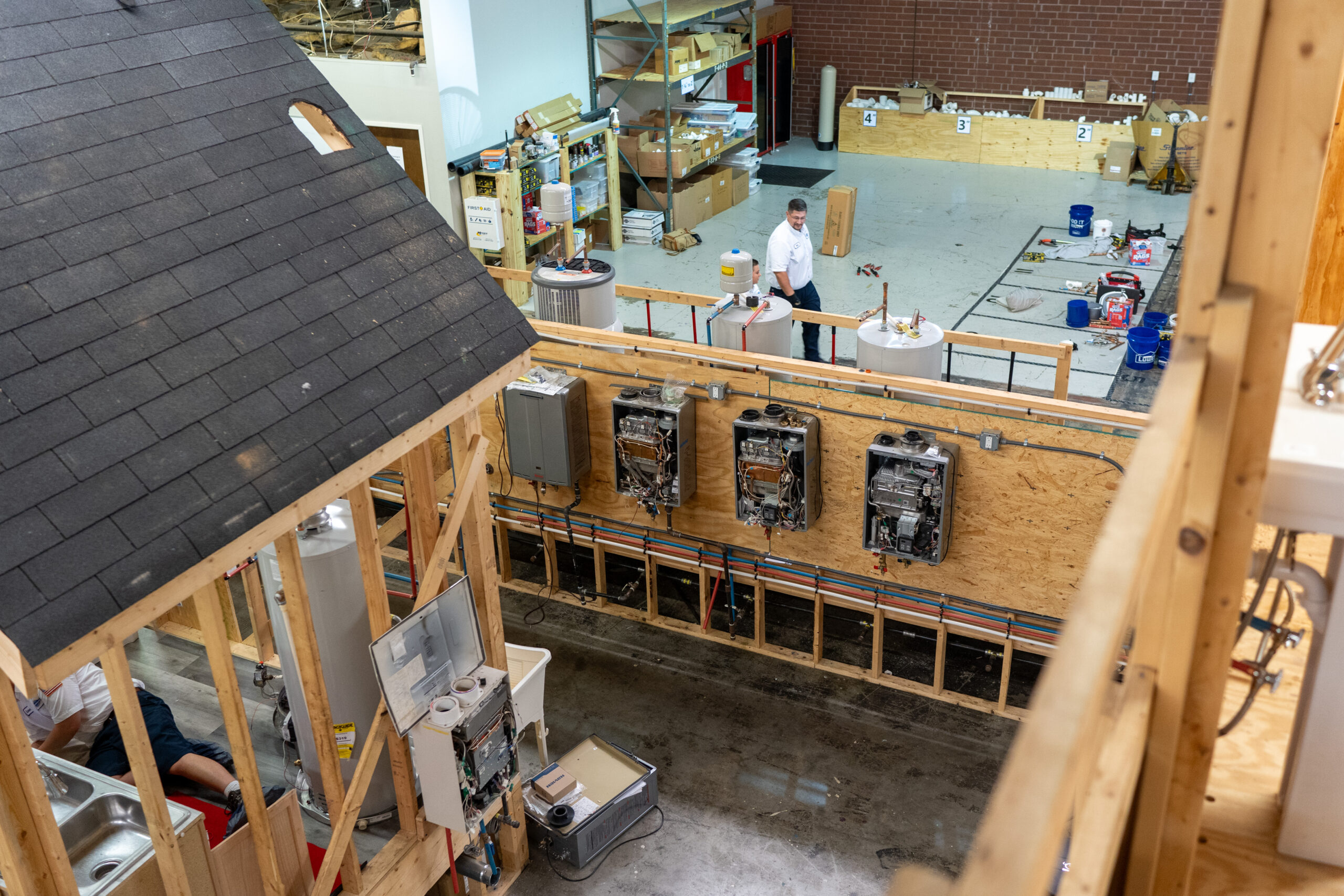 A man works in a large workshop with exposed water heaters, plumbing, and construction materials, next to a partially built structure.