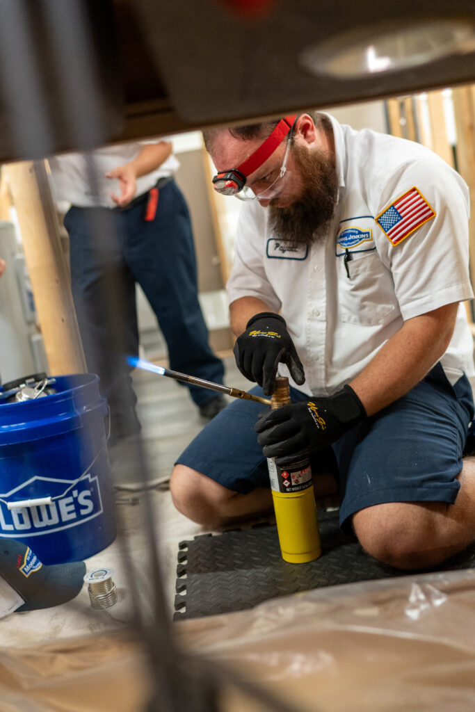 A plumber wearing safety goggles and gloves uses a blowtorch on a pipe next to a blue Lowe's bucket, kneeling on a mat indoors.