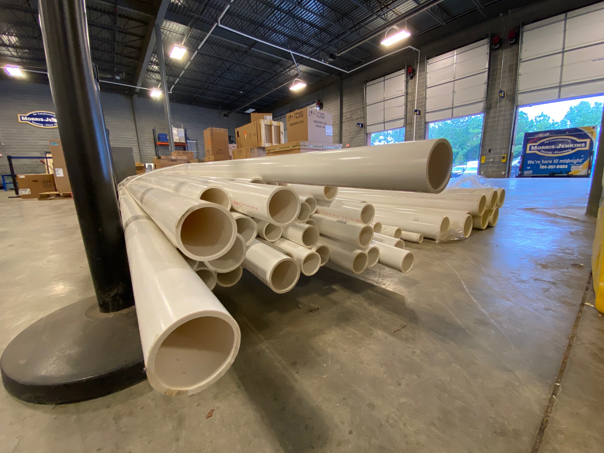 Stacks of white PVC pipes of various diameters are arranged on a warehouse floor, with boxes and equipment visible in the background.