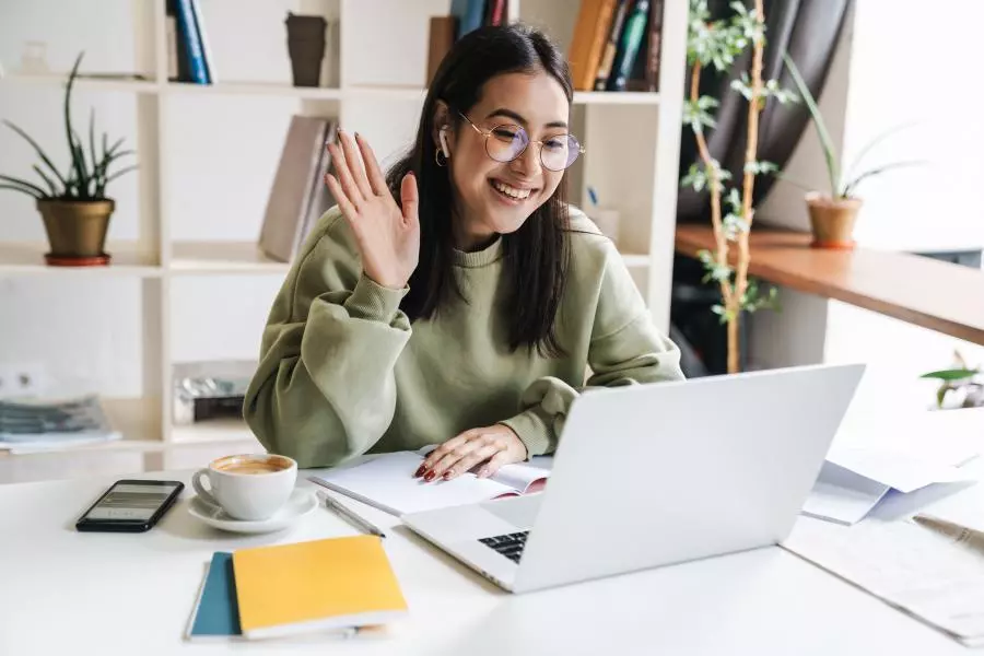 A woman is sitting at a desk with a laptop and waving.