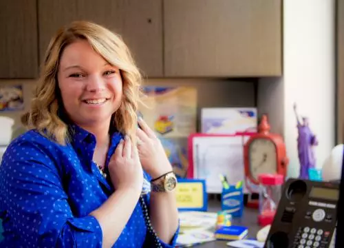 A woman in a blue shirt is smiling in front of a desk.