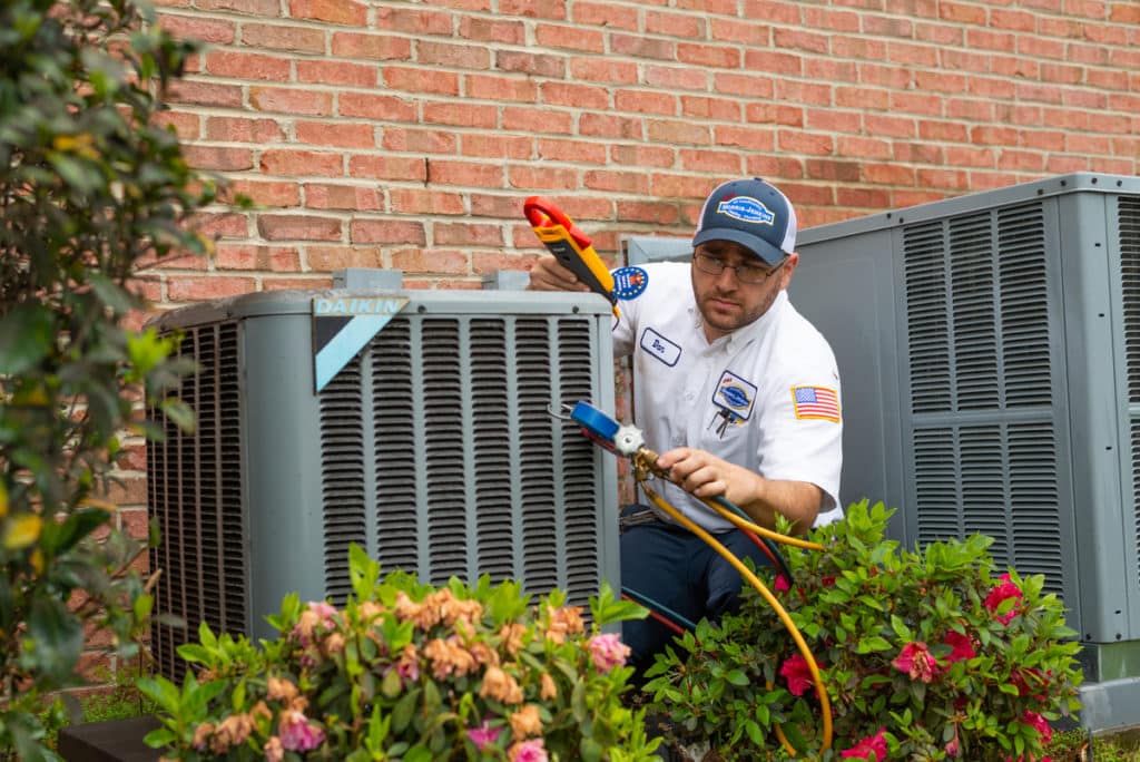 A man fixing an air conditioner in front of bushes.