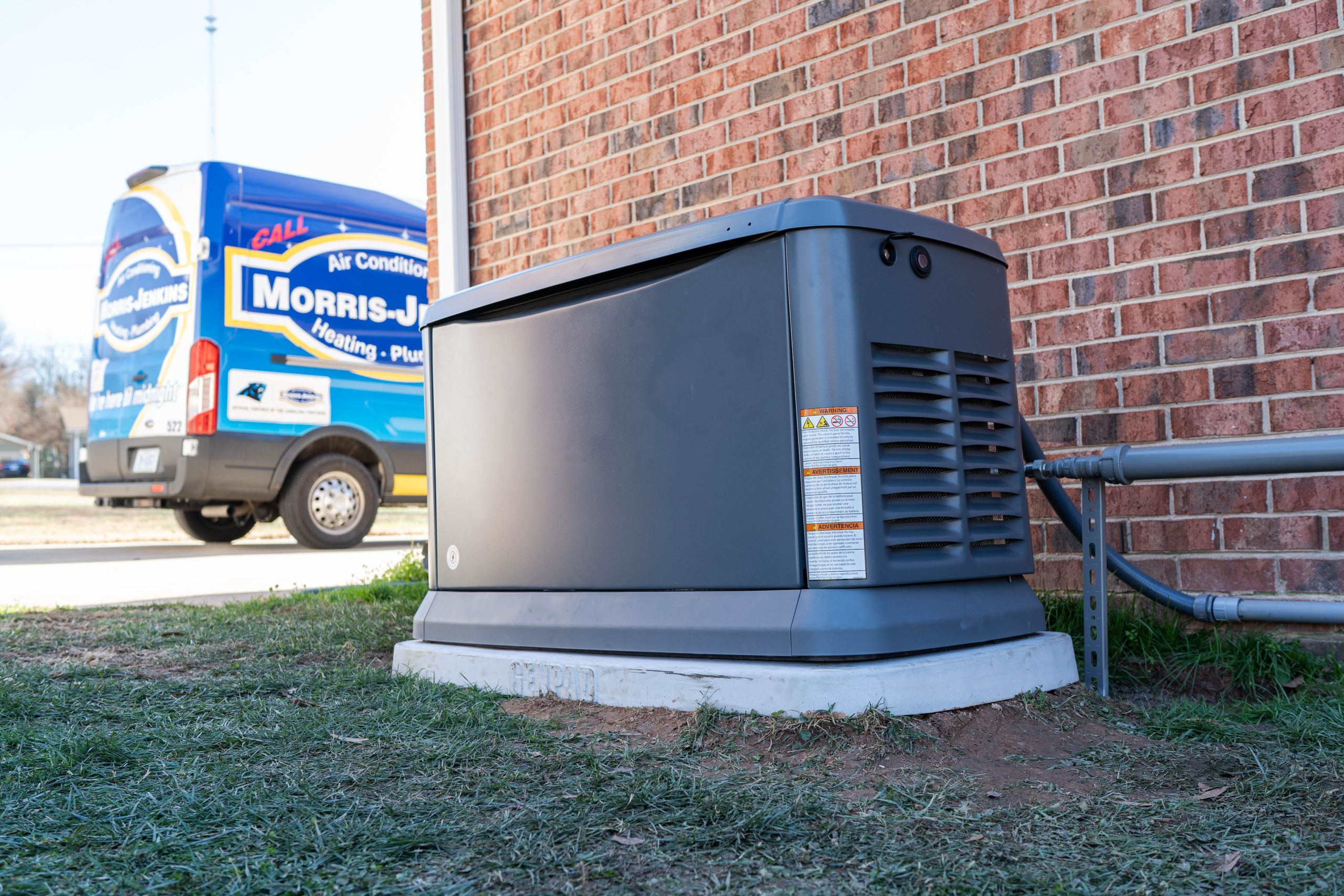 A large, gray backup generator is installed next to a brick building, ready to handle any electrical surge. A service van with "Morris-Jenkins Heating Plumbing Air Conditioning" is parked nearby, ensuring swift support.
