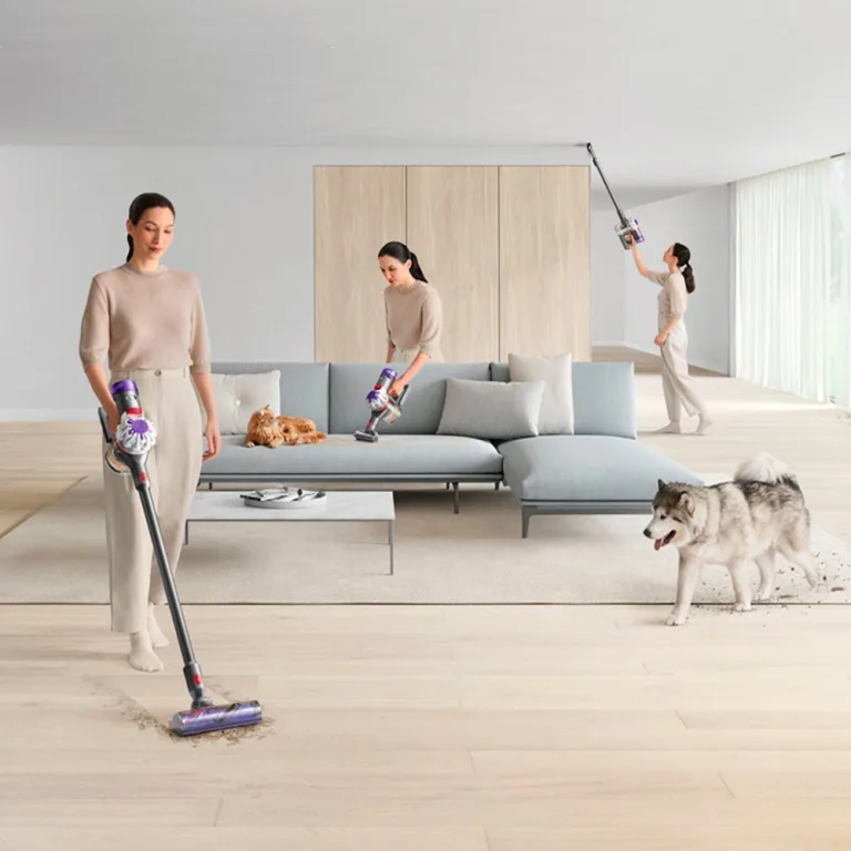 A woman vacuuming the floor with a cordless vacuum cleaner while another woman dusts and a third exercises, with a dog walking by in a modern living room.