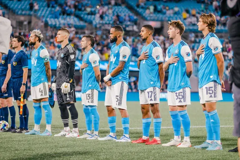 A soccer team in light blue jerseys stands in a line on the field during the national anthem, showing focused expressions.