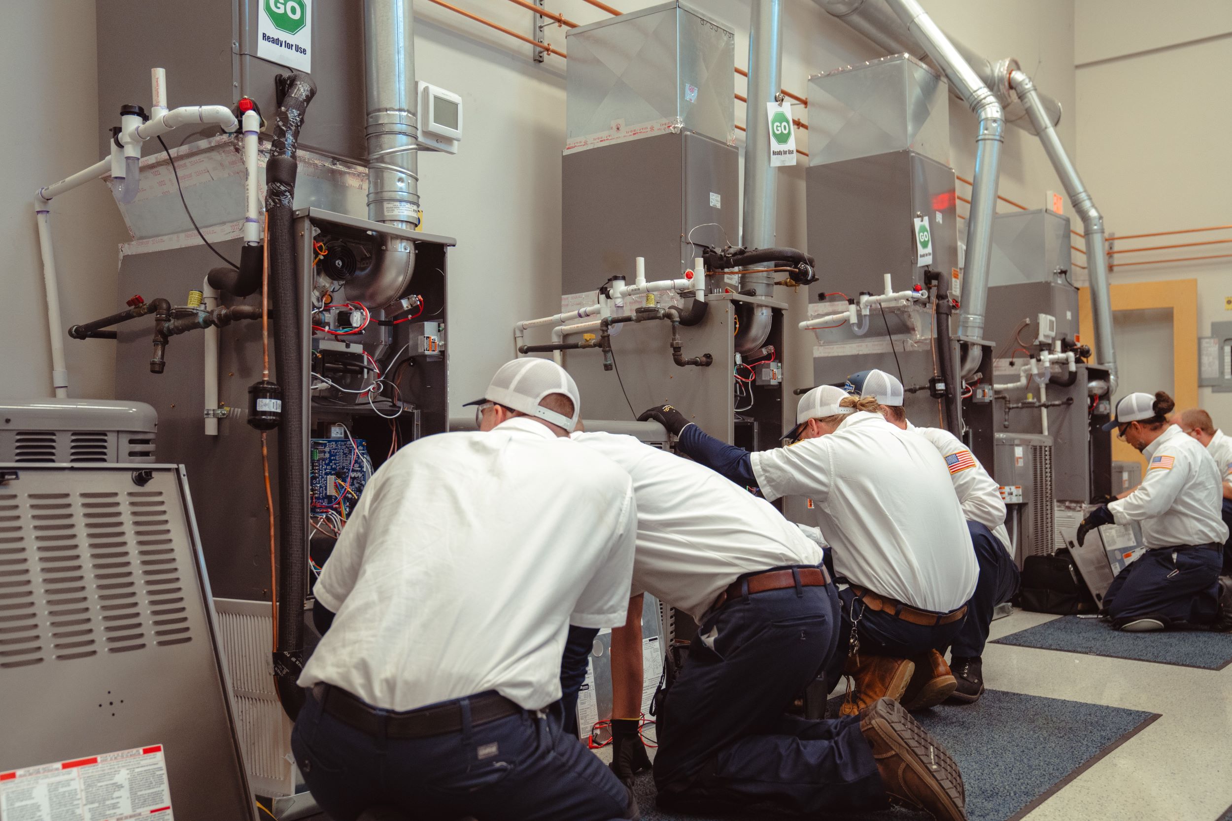 Technicians in white shirts and caps are kneeling and working on large HVAC units lined up in a facility, focusing on maintenance or repair tasks.