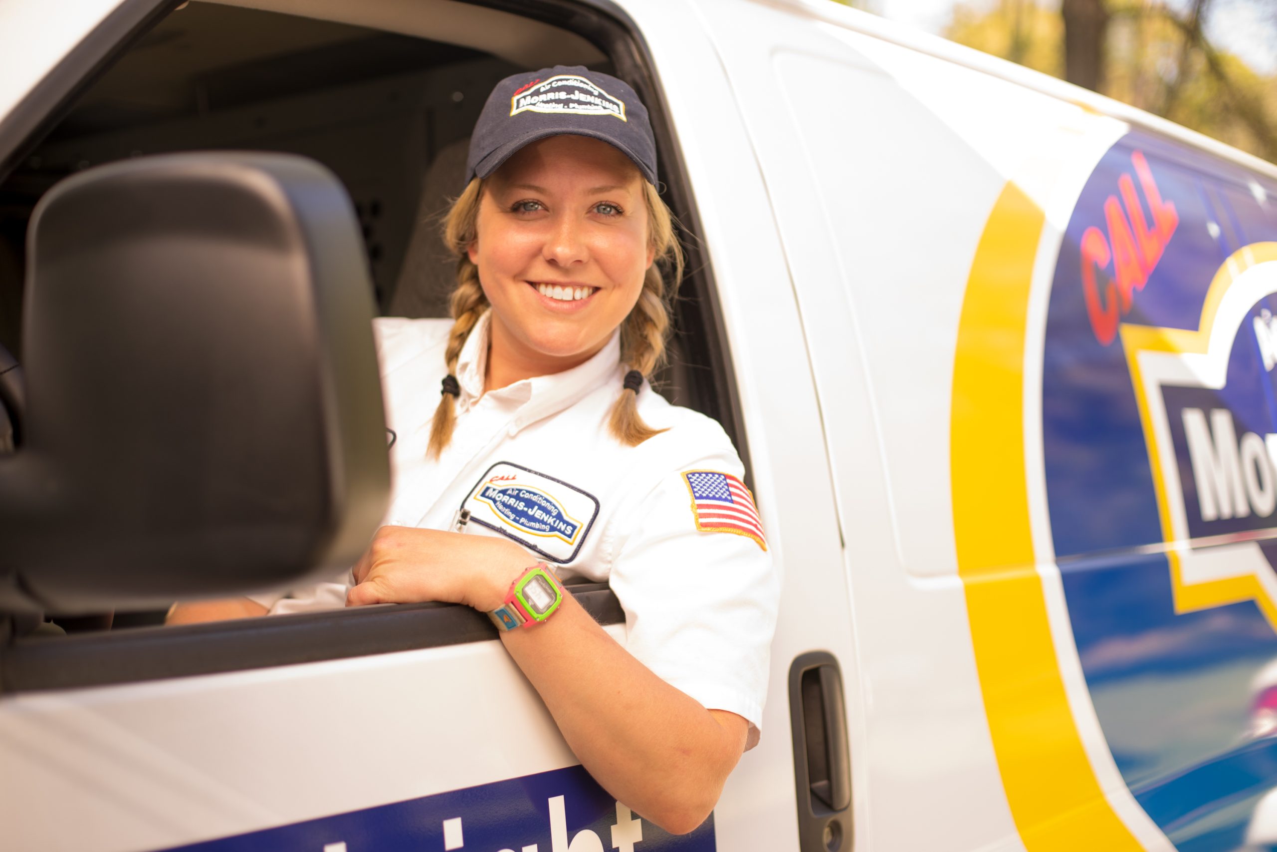 A person in a uniform and cap, with braided hair, smiles from the driver's seat of a service van.