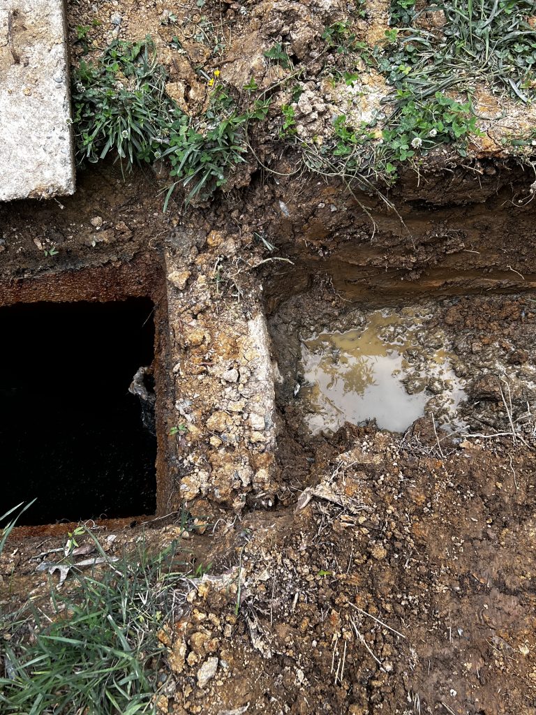 A partially exposed section of ground shows an open concrete manhole next to a muddy hole filled with water. Surrounding the area is grass and soil.