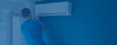 A person in a blue shirt installs a wall-mounted air conditioning unit in a room with blue walls.