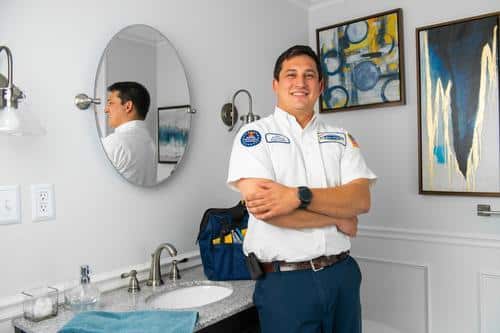 A smiling technician in uniform stands with arms crossed in a bathroom. Tools and a blue towel are on the counter near a sink. Wall art and a round mirror are visible in the background.