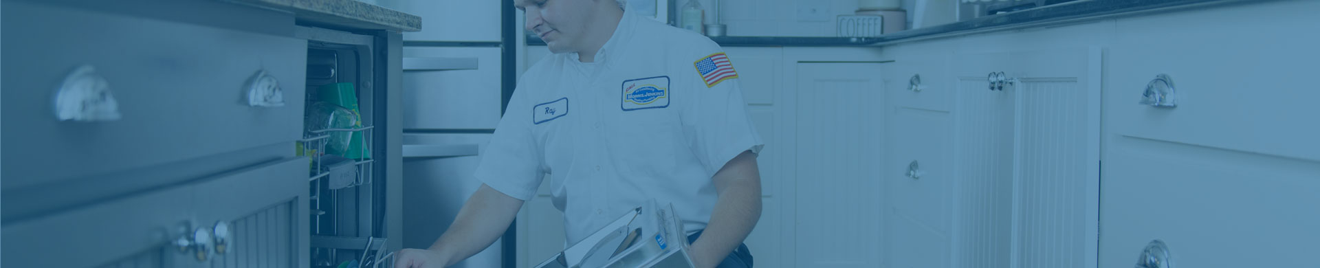 A technician in uniform, with an American flag patch on his sleeve, is repairing or inspecting an appliance in a kitchen setting.