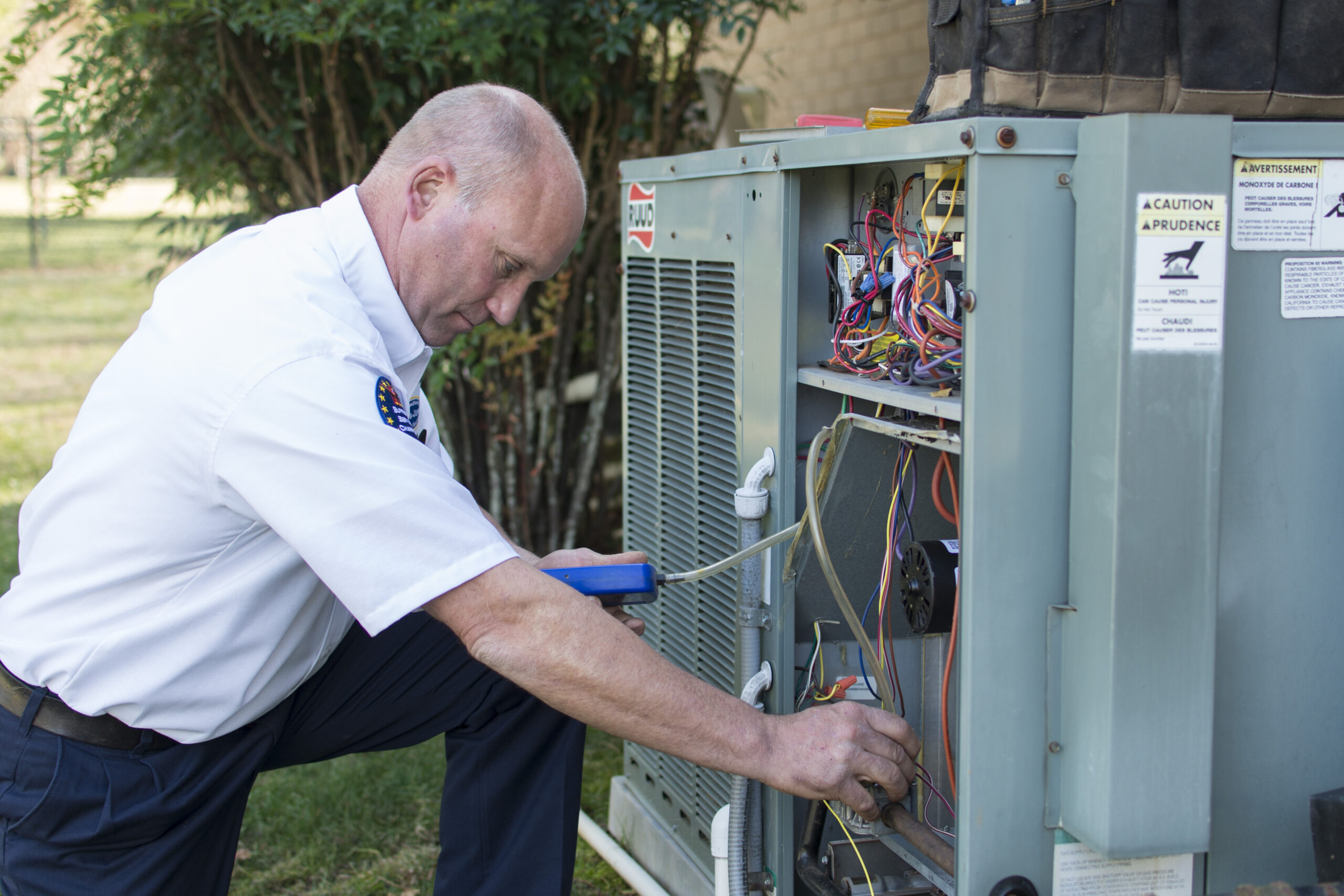 A technician in a white shirt works on the wiring of an outdoor HVAC unit.