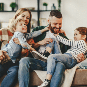 A smiling family of four sits on a couch; the parents hug their two young children, who are laughing and playfully interacting with the father.