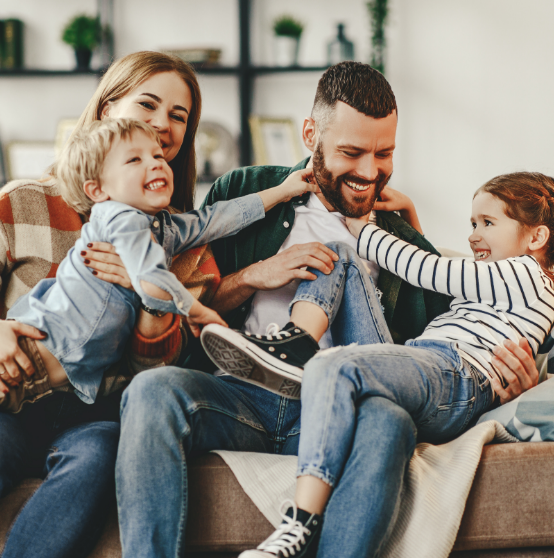 A smiling family of four sits on a couch; the parents hug their two young children, who are laughing and playfully interacting with the father.