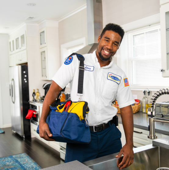 A person in a uniform stands in a kitchen, holding a tool bag and smiling, with a sink and faucet visible in the foreground.