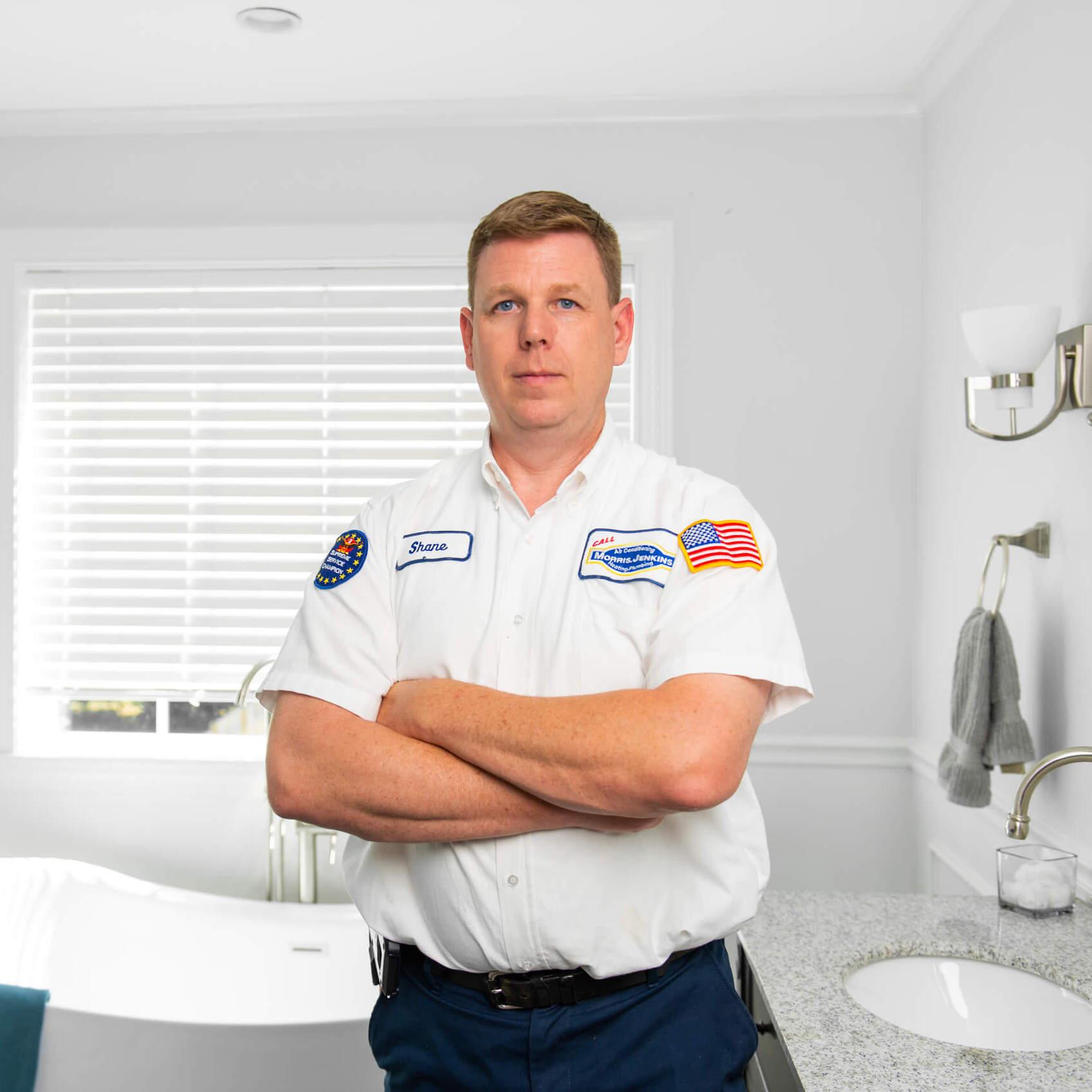 Person in a white uniform with name tag stands in a kitchen by a countertop, smiling. Cabinets and kitchen items are visible in the background.