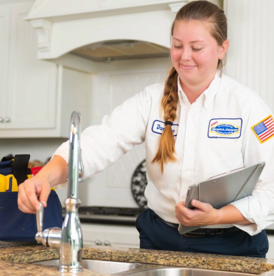 A person in a uniform adjusts a kitchen faucet while holding a tablet. A tool bag sits nearby.