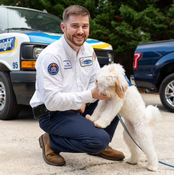 A technician kneels and smiles, petting a white dog standing on its hind legs. A service van and truck are in the background.