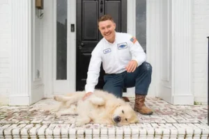 A man in a work uniform kneels on a porch, smiling and petting a golden retriever lying on its side in front of a black door.