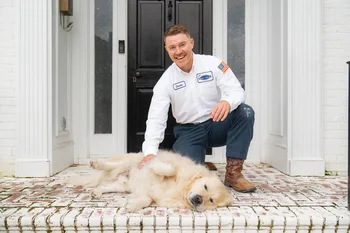 A man in a work uniform kneels on a porch, smiling and petting a golden retriever lying on its side in front of a black door.