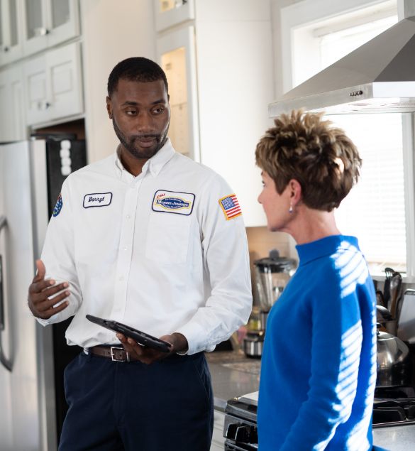 A technician in uniform talks with a woman in a kitchen, holding a tablet and gesturing as they discuss something.