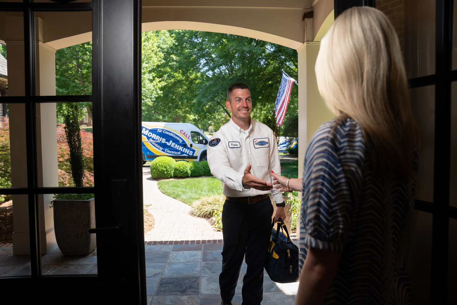A service technician stands at the front door, greeting a woman inside a house. A branded van is parked outside in the driveway.