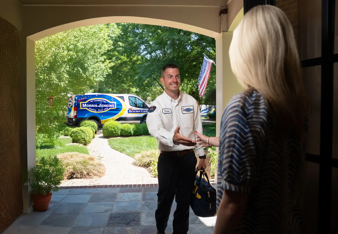 A technician in a Morris-Jenkins uniform shakes hands with a woman at her front door, with a Morris-Jenkins van parked outside on a sunny day.