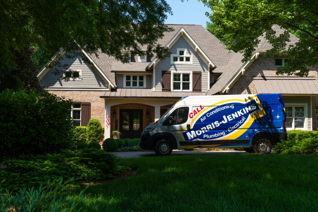 A Moris Jenkins service van is parked in the driveway of a suburban brick house surrounded by trees and greenery on a sunny day.