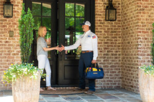 A woman and a uniformed worker shake hands at the front door of a brick house. The worker holds a tool bag and wears a hat and an American flag patch on his sleeve.