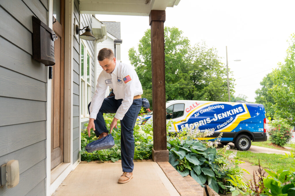 A technician puts on shoe covers before entering a house, with a Harris-Jenkins service van parked in the driveway.