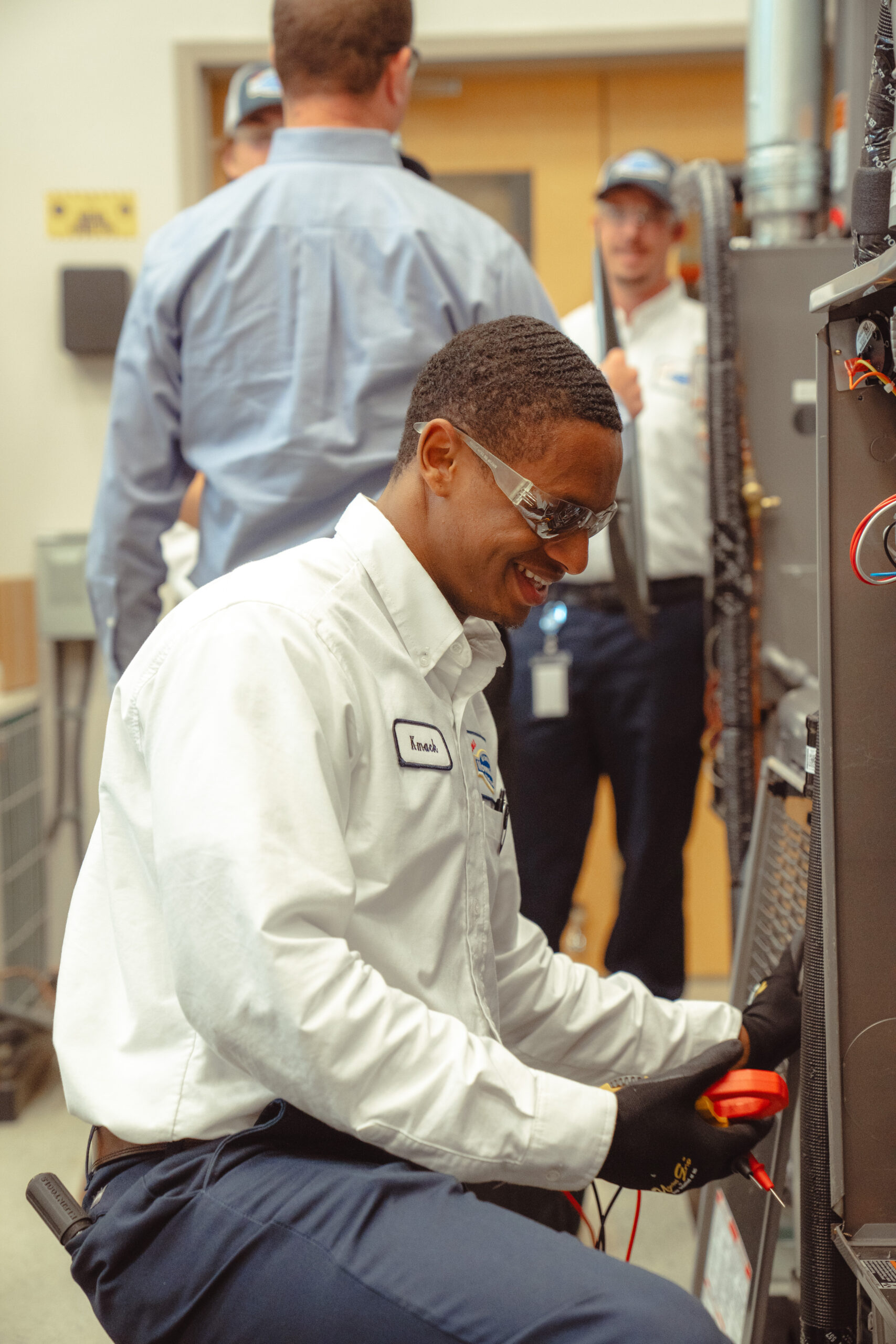 A technician wearing safety glasses and gloves works on electrical equipment while three other people stand in the background in a workshop.