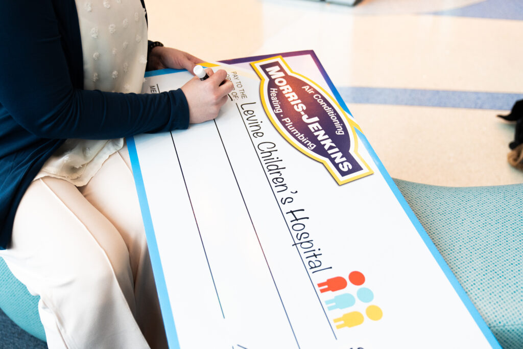 A person writes on a large ceremonial check made out to Levine Children’s Hospital from Morris-Jenkins Heating and Plumbing.