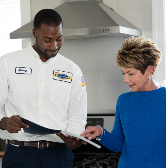 A technician in uniform shows a document to a woman in a blue sweater as they stand in a kitchen.