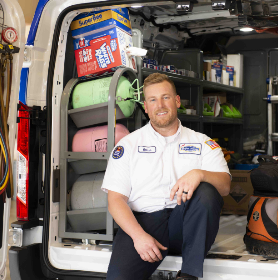 A technician in uniform sits in the back of a work van equipped with various tools and supplies, including refrigerant tanks and cleaning products.