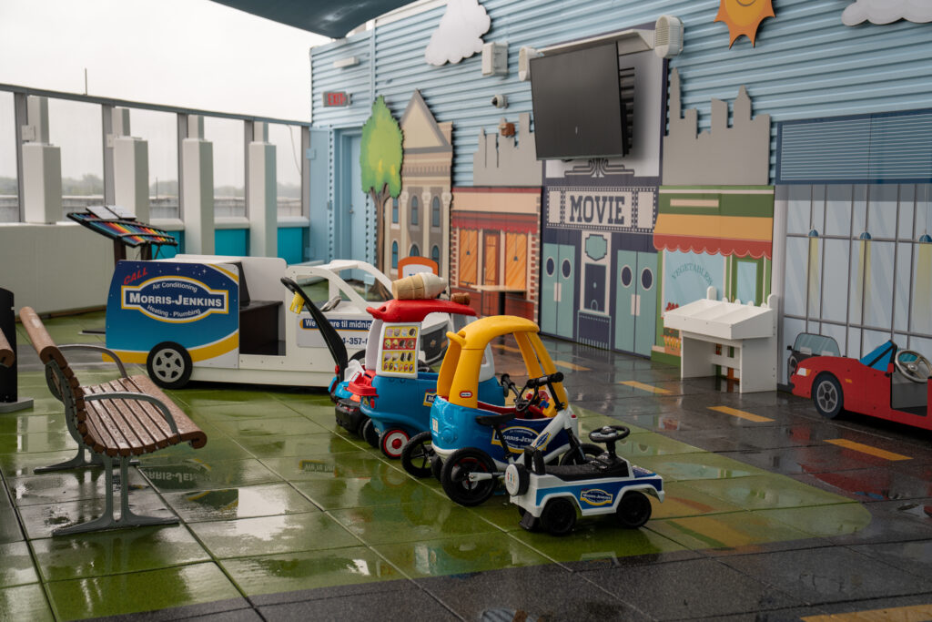 Children's play area with toy cars, a bench, and a movie-themed backdrop on a wet, tiled floor, under a covered outdoor space.