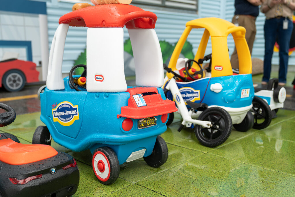 Two toy cars and one tricycle styled with Morris-Jenkins branding are parked on a wet, green surface outdoors. Adults stand in the background.