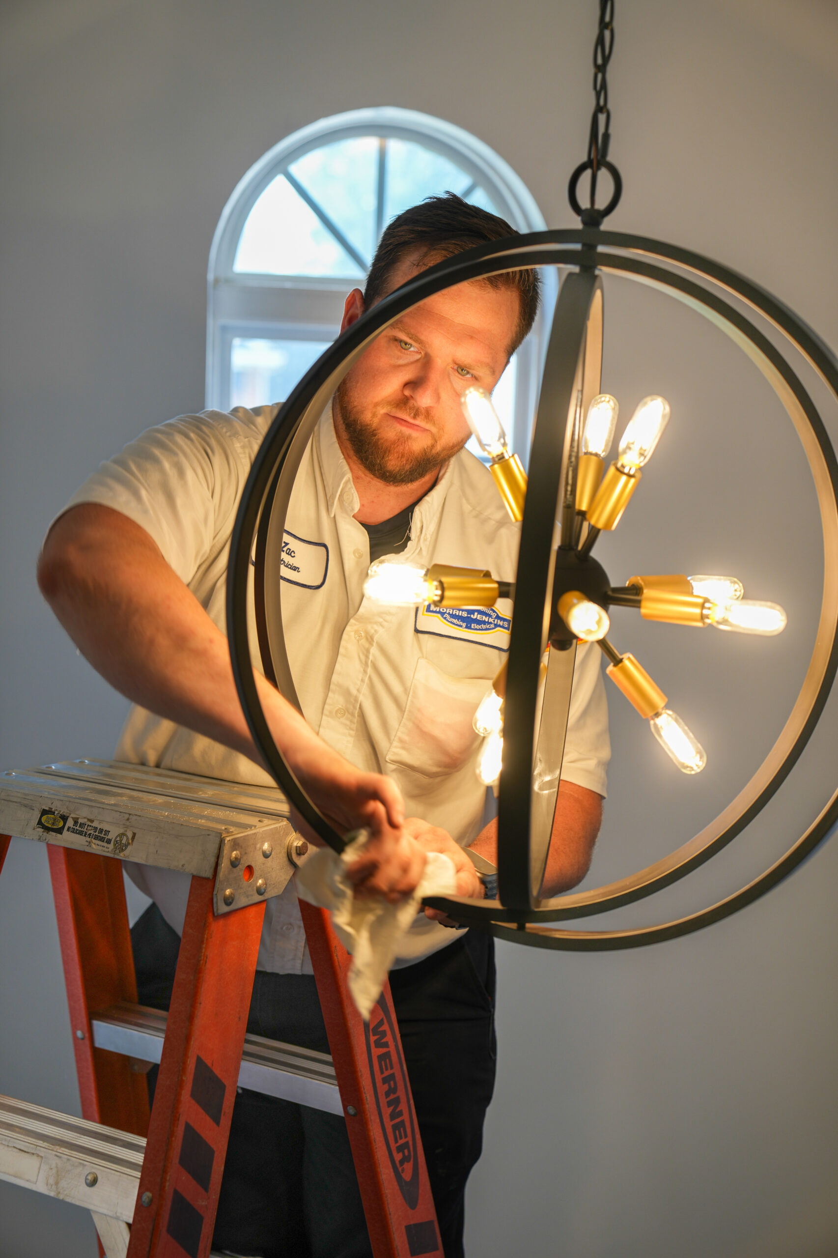 A man standing on a ladder cleans a modern spherical chandelier with lit bulbs in a room with a white wall and arched window.