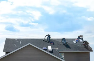 Four workers installing or repairing shingles on the roof of a house under a partly cloudy sky. Roofing materials are scattered across the roof.