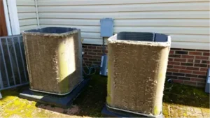 Two outdoor air conditioning units with dirty, clogged condenser coils are positioned on concrete pads next to a building with siding and brick.