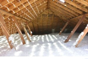 An attic with exposed wooden beams and rafters, partially filled with loose white insulation material on the floor.