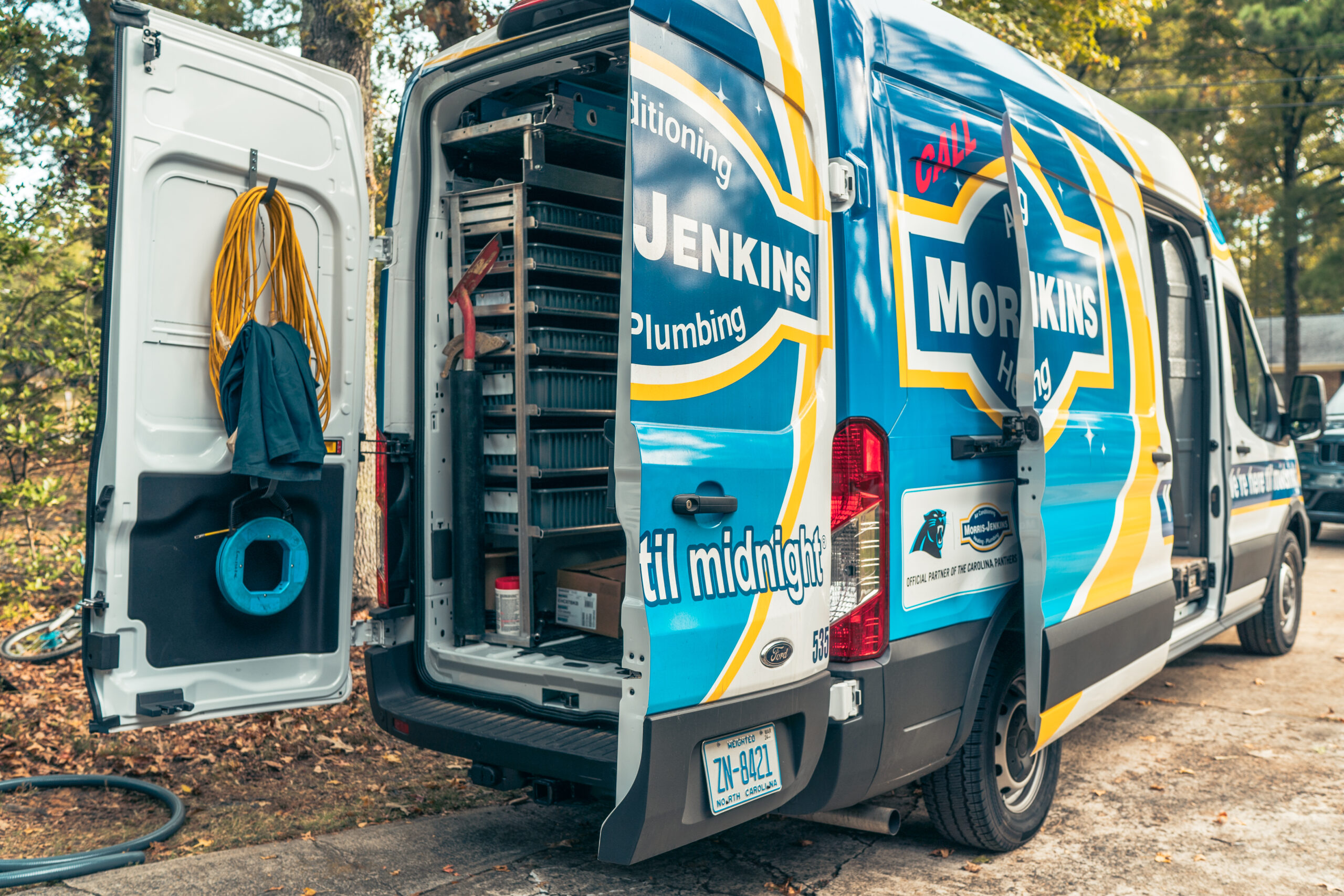 A blue and white service van with its rear doors open, revealing plumbing and HVAC equipment and supplies organized inside.