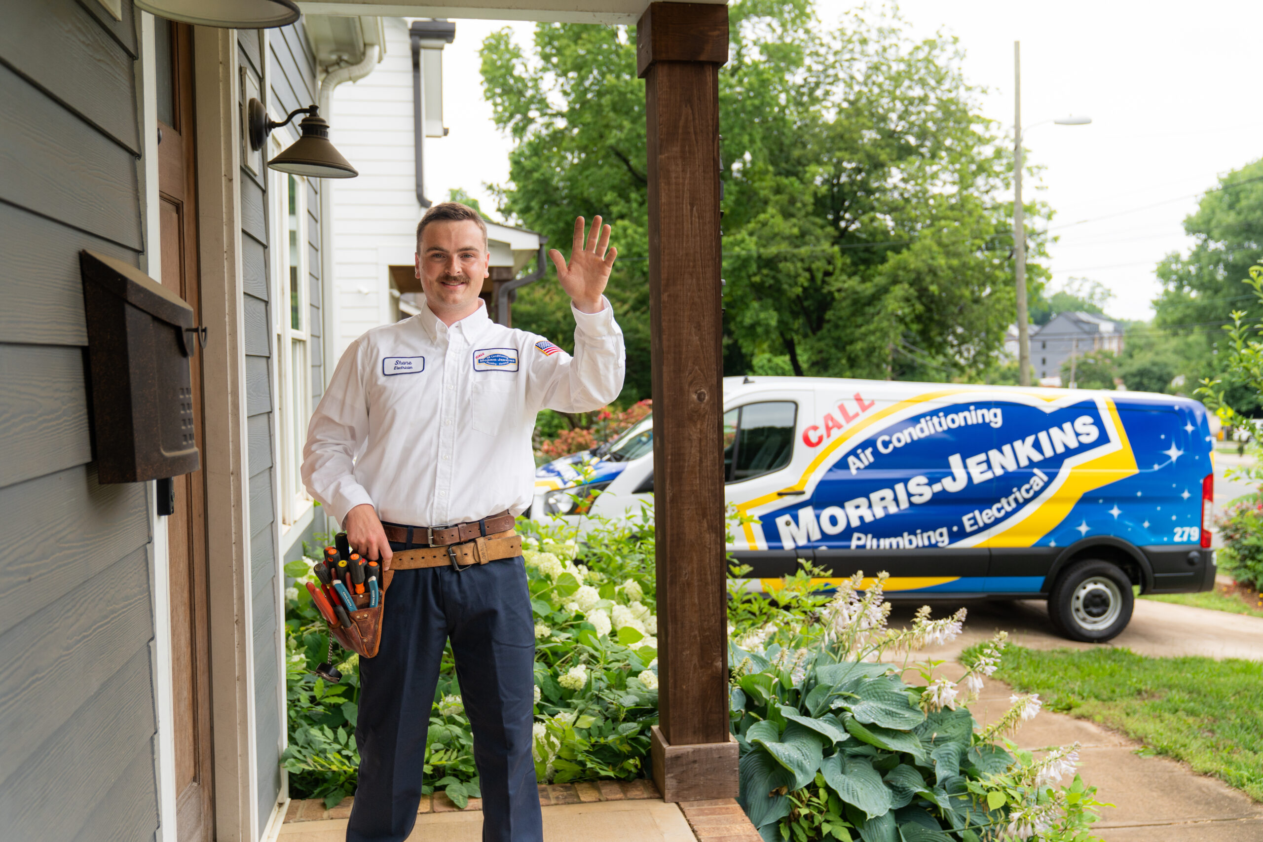 A technician in uniform stands on a porch, waving, with a Morris-Jenkins service van parked in the background among green plants and houses.